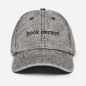 Puede incluir: Gorra de béisbol gris de estilo vintage con las palabras "book person" bordadas en negro. La gorra tiene una visera curva y un botón en la parte superior. La tela tiene un aspecto desgastado y texturizado.