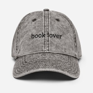 Puede incluir: Gorra de béisbol gris de estilo vintage con el texto "book lover" bordado en negro. La gorra tiene una visera curva y un botón en la parte superior. La tela tiene un aspecto descolorido y desgastado.