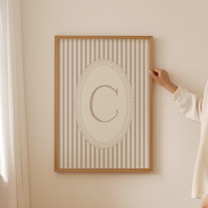 Heirloom-inspired monogram nursery wall art featuring the initial 'C' within a lace frame against classic pink and white stripes.  A natural fit for interiors inspired by the feminine, refined aesthetic of spaces thoughtfully curated by Anthropologie and Studio McGee.