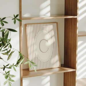 Heirloom-inspired monogram nursery wall art featuring the initial 'C' within a lace frame against classic pink and white stripes.  A natural fit for interiors inspired by the feminine, refined aesthetic of spaces thoughtfully curated by Anthropologie and Studio McGee.