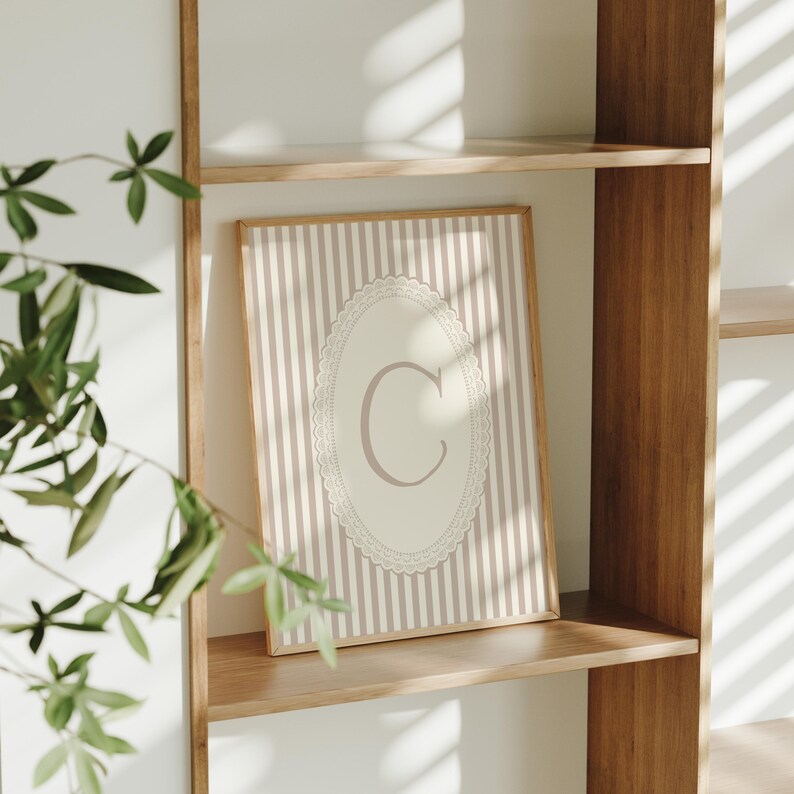 Heirloom-inspired monogram nursery wall art featuring the initial 'C' within a lace frame against classic pink and white stripes.  A natural fit for interiors inspired by the feminine, refined aesthetic of spaces thoughtfully curated by Anthropologie and Studio McGee.