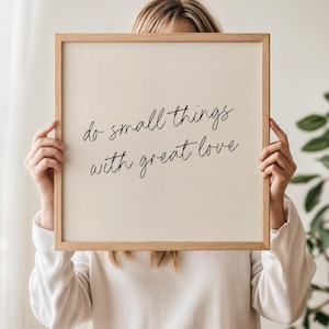 May include: A framed sign with the quote "do small things with great love" in black script. The frame is light brown, and the background is a cream color. The sign is held by a person wearing a white long-sleeved shirt.