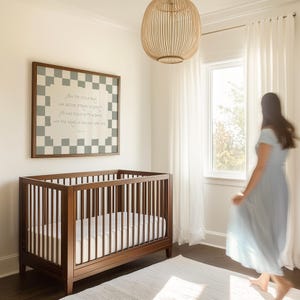 May include: A wooden crib with a white mattress sits in a nursery. A framed artwork with a checkerboard border and text hangs above the crib. A woven light fixture hangs from the ceiling. A woman in a blue dress walks by a window.