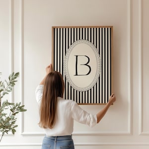 May include: A framed artwork with a black and white striped pattern and a central oval design. The oval displays the letter "B" in black. The frame is a light brown colour.
