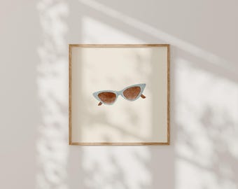 Puede incluir: Una impresión de arte mural cuadrada enmarcada que presenta una ilustración en acuarela de gafas de sol de ojo de gato con monturas azul claro y lentes marrones centradas sobre un fondo color crema sólido, dentro de un marco de madera clara.