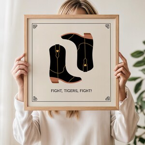 May include: A framed print featuring two black cowboy boots with gold accents and a heart detail. The boots are set against a cream background, with the words "FIGHT, TIGERS, FIGHT!" below.