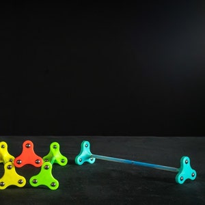 Puede incluir: Un conjunto de cinco accesorios de malabares Flowstox de colores. Los accesorios están hechos de plástico y tienen una forma triangular con tres puntas. Los accesorios son de varios colores, incluyendo amarillo, verde, naranja y azul. Los accesorios están dispuestos en un grupo sobre una superficie negra. El logotipo de Flowstox es visible en la esquina inferior izquierda de la imagen.