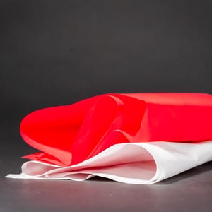 Puede incluir: Un objeto rectangular rojo brillante cubre parcialmente una tela blanca. El objeto es liso y parece estar hecho de un material flexible. La tela blanca está doblada y tiene una textura suave. El logotipo "Juggling Calling" está en la esquina superior izquierda.