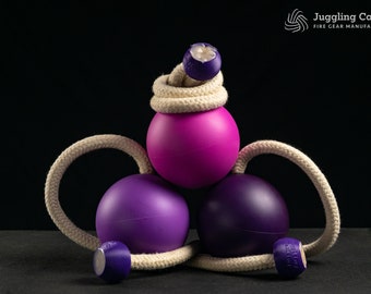 May include: Three juggling balls, one pink and two purple, are connected by a white rope. The balls are arranged in a triangular shape, with the pink ball at the top.