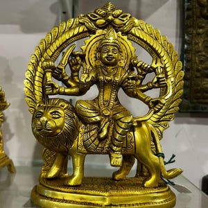 Puede incluir: Una estatua dorada de una deidad sentada sobre un león, con múltiples brazos sosteniendo varios objetos. La estatua es intrincadamente detallada, con un halo y adornos ornamentados. El león tiene una melena detallada y está posicionado sobre una base.