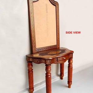 May include: A wooden vanity table with a mirror and intricate inlaid designs. The table has a curved top and three legs. The mirror is framed in wood and has a decorative border. The table is made of dark wood and has a rich, warm color.