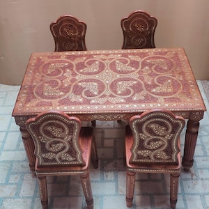 May include: A wooden dining table and four chairs set. The table and chairs are a reddish-brown color with intricate inlaid designs in a lighter color. The table has a rectangular top with ornate patterns.