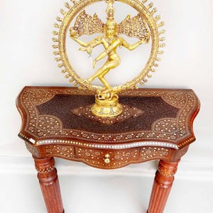 May include: A gold-toned metal sculpture of the Hindu deity Shiva in the Nataraja pose, dancing on a demon, with a halo of hands around him. The sculpture is on a wooden table with intricate inlaid designs.