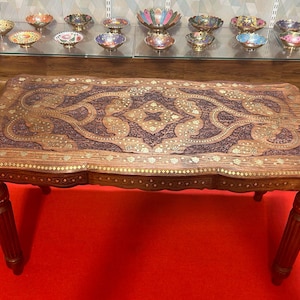 Puede incluir: Una mesa de café de madera oscura con intrincados tallados y detalles dorados. La mesa tiene una tapa rectangular y cuatro patas.