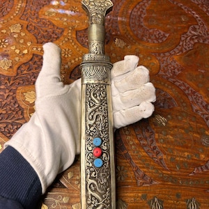 May include: An ornate dagger with a silver-colored sheath and handle. The sheath is decorated with dragon motifs and inlaid with red and blue stones. The handle has intricate floral patterns. The dagger is displayed on a wooden surface.