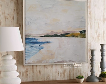 May include: A framed abstract landscape painting with a beach scene. The artwork features a white sandy beach, blue water, and green and yellow hills under a cloudy sky. The painting is in a white frame and is displayed on a wooden surface.