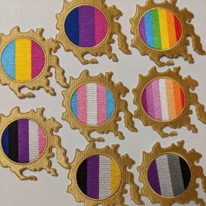 May include: Embroidered patches featuring various LGBTQ+ pride flags, including rainbow, transgender, bisexual, pansexual, non-binary, asexual, and genderqueer designs. Each patch has a gold-colored, ornate border and a circular center.