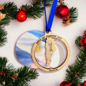 May include: A wooden Christmas ornament with a blue ribbon, featuring the Argentina flag and a map outline with the text "WORLD CUP CHAMPIONS 2022". The ornament is surrounded by evergreen branches and red ornaments.