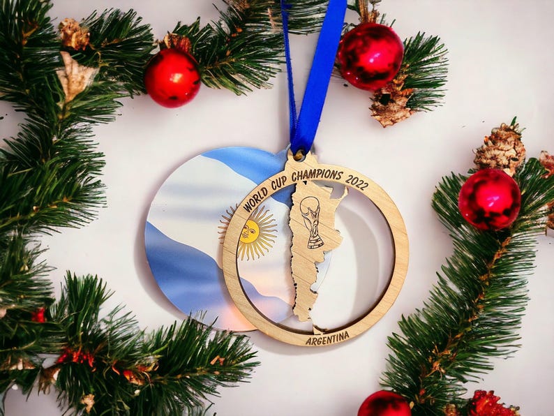 May include: A wooden Christmas ornament with a blue ribbon, featuring the Argentina flag and a map outline with the text "WORLD CUP CHAMPIONS 2022". The ornament is surrounded by evergreen branches and red ornaments.