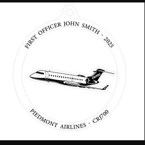 May include: A white, circular ornament featuring a black and white illustration of a CRJ700 aircraft. The text reads "FIRST OFFICER JOHN SMITH - 2025" and "PIEDMONT AIRLINES - CRJ700". The ornament has a small hole at the top for hanging.