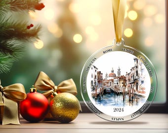 May include: A Christmas ornament with a watercolor painting of a canal scene, featuring buildings and a bridge. The ornament is round with the words "Rome", "Venice", "Cinque Terre", "Pisa", "Amalfi Coast", "Naples", "Taormina", "Palermo", and "Siracusa" around the edge. The year "2024" is printed on the ornament. Red and gold Christmas ornaments and a gold bow are in the foreground.