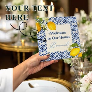 May include: A decorative sign with a blue and white tile pattern border, featuring lemons and the text "Welcome to Our House Benvenuti". The sign is held in front of a table setting with candles and flowers.