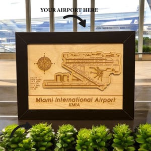 May include: A framed wooden map of Miami International Airport, KMIA, with a detailed laser-cut design. The map includes a compass rose and airport information. The frame is black, and the background is light wood.