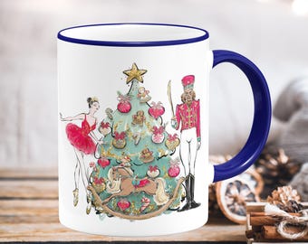 May include: White ceramic mug with a blue handle and rim, featuring a Christmas-themed illustration. The design includes a decorated Christmas tree, a ballerina in a red tutu, and a nutcracker soldier. The mug is decorated with gold accents.