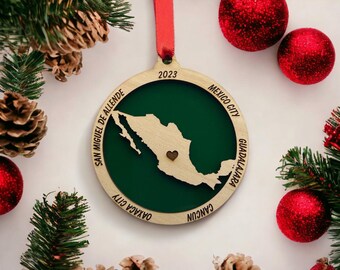 May include: A wooden Christmas decoration depicting a map of Mexico, with a heart detail. The ornament is set against a backdrop of red baubles, pine cones, and foliage. The year 2023 is shown at the top, with city names around the edge.