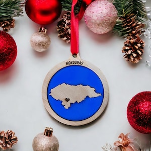 May include: A wooden Christmas ornament featuring the map of Honduras, set against a blue background. The word "HONDURAS" is printed at the top. The ornament is surrounded by red and gold Christmas balls, pine cones, and evergreen branches.