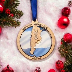 May include: Wooden Christmas ornament with a blue ribbon, featuring the Argentina flag and a trophy. The ornament is surrounded by red Christmas balls and green pine branches, creating a festive holiday scene.