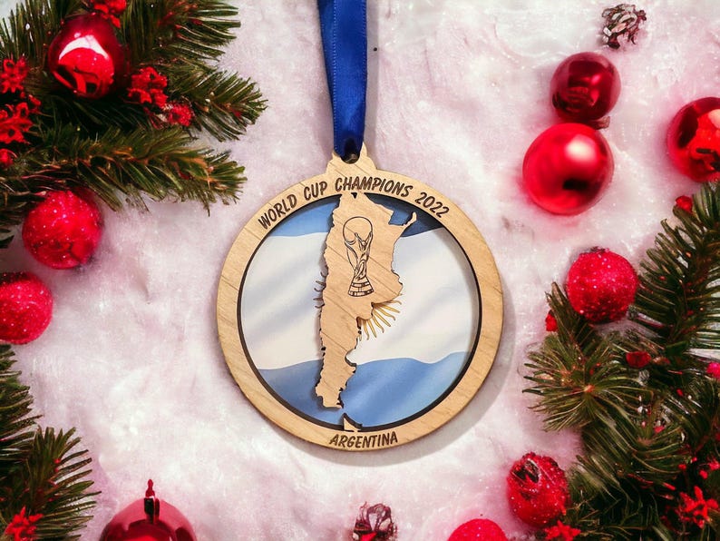 May include: Wooden Christmas ornament with a blue ribbon, featuring the Argentina flag and a trophy. The ornament is surrounded by red Christmas balls and green pine branches, creating a festive holiday scene.