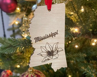 May include: A wooden ornament in the shape of the state of Mississippi, with the state name and a magnolia flower engraved. The ornament hangs from a red ribbon and is displayed on a Christmas tree with other ornaments.