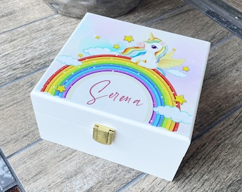 Può includere: Scatola di legno bianca con coperchio incernierato e chiusura color oro. Il coperchio presenta un'illustrazione colorata di un unicorno, arcobaleno, nuvole e stelle. Il nome "Serena" è scritto in corsivo sul coperchio.