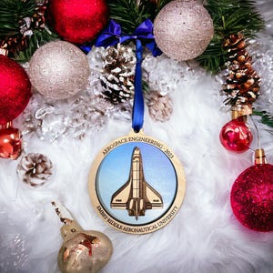 May include: A wooden Christmas ornament featuring a space shuttle design, with the text "AEROSPACE ENGINEERING - 2023" and "EMBRY-RIDDLE AERONAUTICAL UNIVERSITY". The ornament hangs with a blue ribbon among red and silver Christmas ornaments.