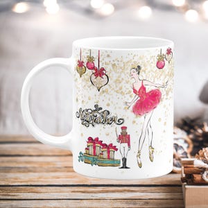 May include: White ceramic mug featuring a ballerina in a pink tutu and the words "The Nutcracker." The mug also has illustrations of wrapped gifts, ornaments, and a toy soldier. Gold glitter accents the design.