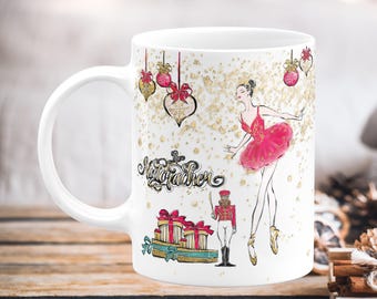 May include: White ceramic mug featuring a ballerina in a pink tutu and the words "The Nutcracker." The mug also has illustrations of wrapped gifts, ornaments, and a toy soldier. Gold glitter accents the design.