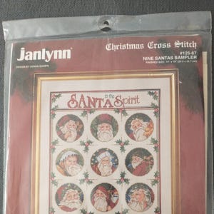 May include: A Christmas cross-stitch kit featuring a sampler design with nine different Santa Claus illustrations. The text reads "in the Santa Spirit" and "Christmas". The finished size is 14 x 18 inches.