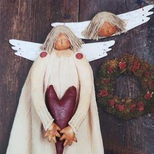 May include: A handcrafted angel doll with a cream-colored dress, holding a burgundy heart. The angel has a straw-like hair and painted facial features. A small angel head and a wreath with red roses are also visible.