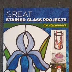 May include: A book cover titled "GREAT STAINED GLASS PROJECTS for Beginners" features a blue iris design and examples of stained glass projects. The cover is predominantly blue with white text and illustrations, showcasing various stained glass techniques.