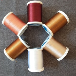 May include: Six spools of thread in a circle. The spools are in various shades of brown, beige, and white. The spools are arranged in a circle with the white spool at the bottom.