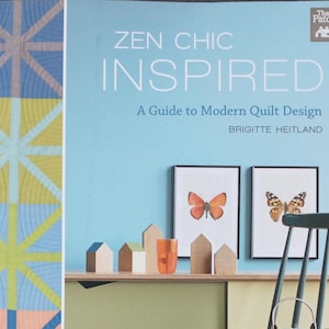 May include: A softcover book titled Zen Chic Inspired: A Guide to Modern Quilt Design by Brigitte Heitland. The cover features a light blue background, a geometric quilt pattern on the left, and a shelf with wooden house blocks and butterfly art.