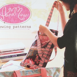 May include: A woman holds a colorful fabric tote bag with the text "Anna Maria sewing patterns" on the front. The bag has a floral print and pink and white stripes.