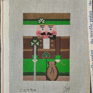 May include: Needlepoint canvas featuring a St. Patrick's Day themed nutcracker. The design includes a brown and green uniform, a shamrock, and a sack of gold. The text "the needle point" is printed on the canvas border.