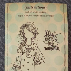 May include: A light green and cream-colored stamp with a drawing of a woman holding a mug. The stamp has the words "Stay Cozy & Feel the Warmth" in a handwritten font. Instructions are printed at the top.