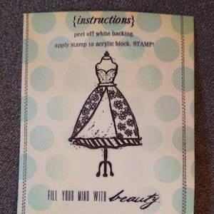 May include: A light green card with instructions for a stamp. The card features a black and white illustration of a dress on a stand. The text reads "{instructions}" and "FILL YOUR MIND WITH beauty."