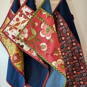 May include: Three colorful fabric aprons with different patterns. The aprons are made of denim, red floral fabric, and a white fabric with red and yellow floral designs. The aprons have red and white polka dot trim.