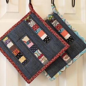 May include: Two square denim pot holders with colorful patchwork accents. The pot holders have a dark blue denim base with rectangular patchwork designs in various colors and patterns. The edges are trimmed with a red polka dot fabric and a blue and white patterned fabric.