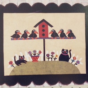 May include: A black and white felt applique wall hanging with a birdhouse, five black birds, two black cats, and a flower garden. The background is a light beige color and the frame is black with a scalloped edge.