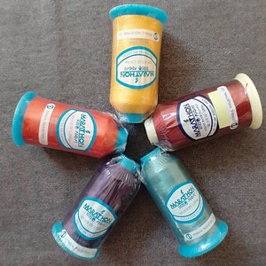 May include: Five spools of thread in different colors, including gold, red, purple, green, and brown. Each spool has a label that says "Marathon" and "Made in Korea".